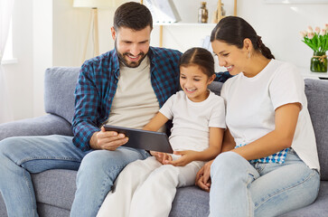 Happy family using tablet together, father and mother with daughter sitting on sofa at home. Parents and child browsing and reading news in social media, looking at screen and smiling in living room