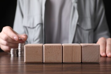 Woman stacking coins at wooden table, focus on blank cubes. Mockup for design