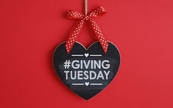 Black heart shaped chalkboard sign with white text # GIVING TUESDAY hanging by a red polka dot ribbon against a bright vibrant background for charity fundraising