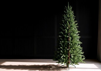 Tall slender artificial christmas tree with dense green needles stands in a room with dark background and sunlight casting shadows