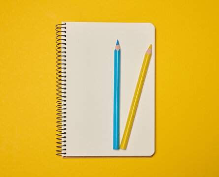 A spiral-bound notebook with blank pages, topped with blue and yellow pencils, sits against a yellow background. - Powered by Adobe