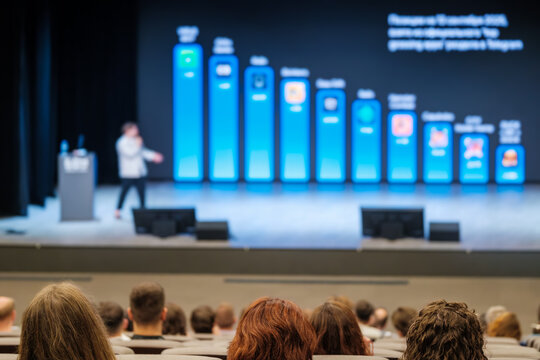 Blurry stage presentation with presenter, row of large blue callout panels and audience silhouettes indoors