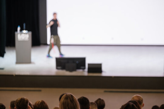 Presenter on stage delivers talk to attentive crowd with podium and bright backdrop stage lights focus