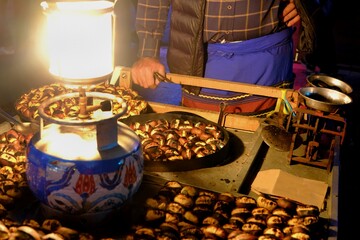 A view of chestnut kebab, a traditional Turkish street food, being cooked. A picnic lamp illuminates the stall. The vendor roasts chestnuts over a charcoal fire using tongs.
