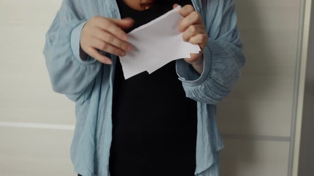 Close up of woman in light blue shirt ripping white A4 paper sheet into multiple pieces with both hands.