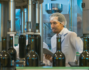 Two inspector wearing lab coat and hairnet check production detail on tablet in modern food manufacturing facility. Machinery and conveyor belt in background reflect hygiene and industrial standard.