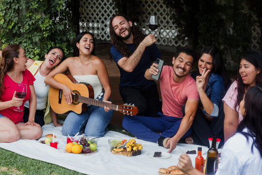 group of latin young adults friends and woman playing guitar sitting on picnic blanket in sunny day in Mexico Latin America. Hispanic diverse and multi generation people in outdoors activities - Powered by Adobe