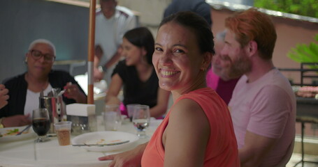 Woman smiling brightly at the camera during a lively family BBQ lunch, surrounded by friends and family enjoying an outdoor meal in a warm and cheerful atmosphere