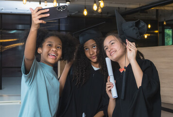 Friend take joyful selfie during graduation ceremony. graduation gown cap, holding diploma scroll, indoor setting feature warm lighting decorative hanging bulb, creating cozy celebratory atmosphere.