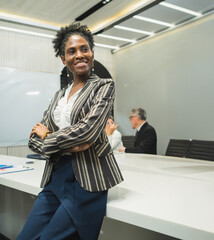 professional woman stand confident arm cross in modern office conveyed strong professional presence. background include colleague working at desk laptop visible reflect  contemporary workspace.