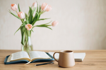 Open Bible on a wooden table and ceramic coffee mug and a vase with tulips.