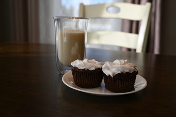 Two black bean and chocolate muffins with coconut whipped cream for a healthier snack on a small white plate and a brown tabletop with warm plant based, no dairy milk in a glass. 