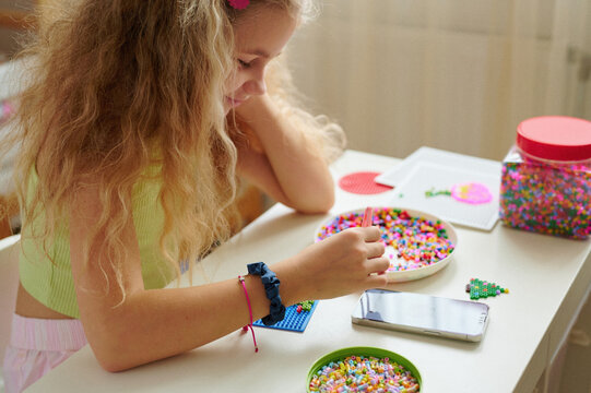 girl engaged in bead crafting enjoys colorful creativity. young artist focused on vibrant beadwork with smartphone on table. child exploring fun diy arts at home, cultivating creativity