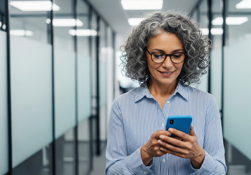 Smiling middle aged woman with curly gray hair wearing glasses and a blue striped shirt walks down a modern office hallway while looking at her smartphone