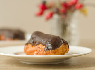 A chocolate eclair on a plate, with a vase of red autumn berries in the background
