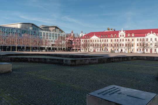 Modern glass facades of the DomQuartier meet the colorful Green Citadel with its golden domes on Magdeburg&rsquo;s Domplatz under a clear blue sky.