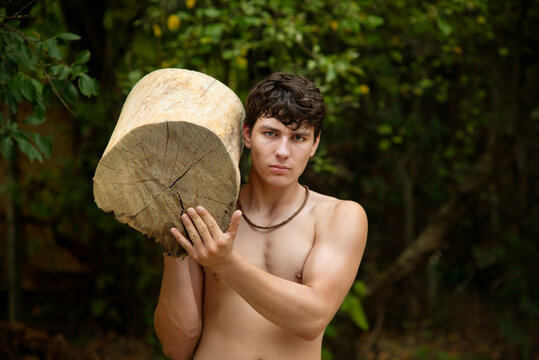 A guy is doing sports outdoors, lifting heavy wooden stumps, squatting and doing push-ups. A shirtless young man stands in a wooded area, cradling thick, freshly cut tree trunk. Young athlete