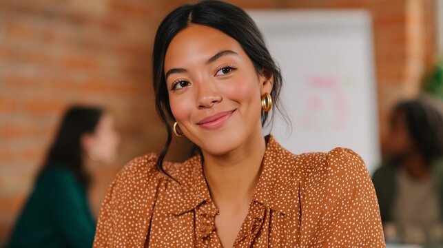 Latina woman wearing an orange brown polka dot blouse smiles warmly while seated at a table, with a blurred background of colleagues engaged in conversation