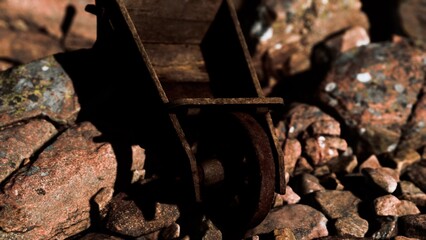 A weathered wheelbarrow made of rusted metal rests among a bed of smooth, colorful stones. The sun shines softly, highlighting the textures and colors in this tranquil garden spot.