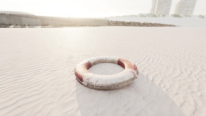 A lifebuoy sits on a tranquil sandy beach, gently illuminated by the soft rays of the sunrise. Nearby buildings stand in the distance, adding a serene backdrop to the peaceful scenery.