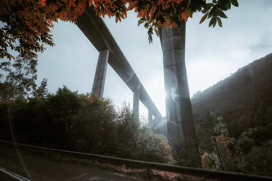 Modern concrete viaduct rising above misty forest hills of Asturias during rain, sunlight flares breaking through clouds on the Camino de Santiago pilgrimage route, dramatic autumn travel scenery
