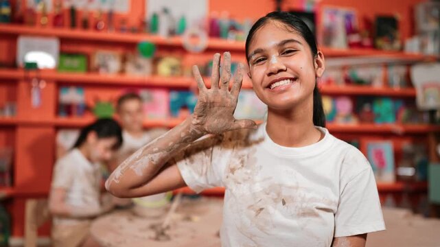 Happy caucasian girl pose at camera while diverse children modeling clay behind. Cute student wearing dirty shirt while looking at camera at workshop in art lesson. Blurring background. Edification