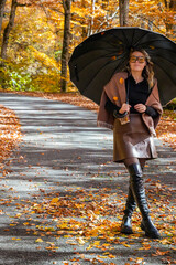 Obraz premium Woman Standing on Autumn Road Holding Black Umbrella, Looking at Camera