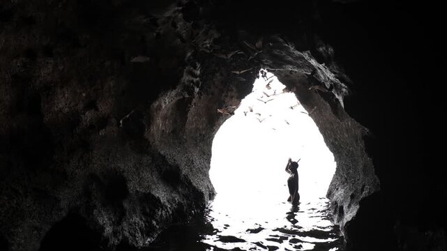 A female stands waist-deep in a cave, silhouetted against bright backlight, framed by the cavern walls as countless bats fly overhead, creating a dramatic, wild, and cinematic natural scene.