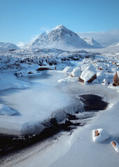 Bright, freezing winter scene in Glencoe featuring the iconic Buachaille Etive Mor rising above the River Etive. The frozen river and snow-covered rocks dominate the foreground under a clear blue sky