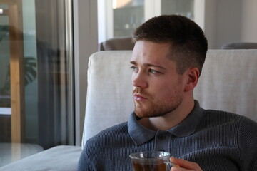 sick young man with cold sitting on couch drinking tea while looking out window at home