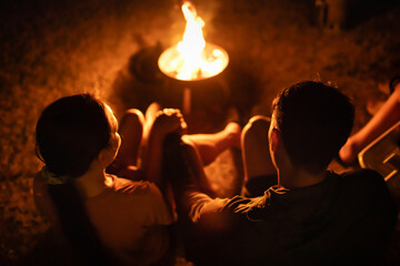 Two people sitting by a warm campfire at night, enjoying a cozy outdoor moment. Represents camping,...