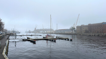 Fototapeta premium Foggy harbor scene with calm water, empty docks, a single moored sailboat, cranes, and industrial buildings in the background, creating a serene waterfront atmosphere.