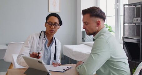 Doctor consults man patient in medical clinic using tablet. Physician explains consultation and discusses symptoms, diagnosis, treatment, and appointment. Healthcare and medicine concept. - Powered by Adobe