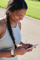 Smiling african millennial woman using smartphone in a park after training, wearing sportswear and smartwatch. Close up side view conveys mobile phone connection, positivity healthy active lifestyle.