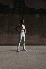 Confident african millennial woman in sportswear standing outdoors by a concrete wall after training. Natural sunlight and long shadow create a minimalist portrait conveying discipline and strength.