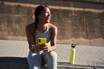 Smiling african millennial woman checking smartphone during a break in the park after training, wearing sportswear and smartwatch. Side view close up conveys connection, positivity and healthy living.