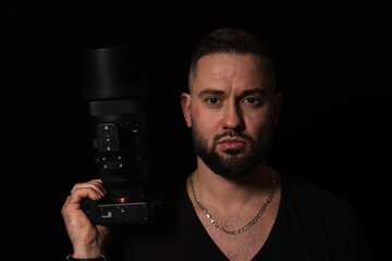 Studio portrait of a serious bearded man holding a professional digital camera with a large lens on a black background.