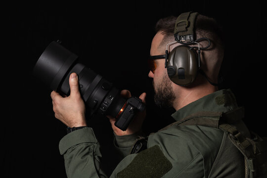 Tactical photographer or videographer in full gear holding a professional camera with a large lens against a black background. - Powered by Adobe