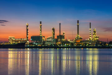 Obraz premium Oil refinery plant illuminated at twilight with bright lights reflecting on water. Industrial landscape showing petrochemical complex, distillation towers, smokestacks, and infrastructure.