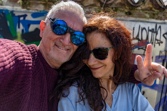 Happy senior couple embracing, wearing sunglasses, and making a peace sign in front of graffiti