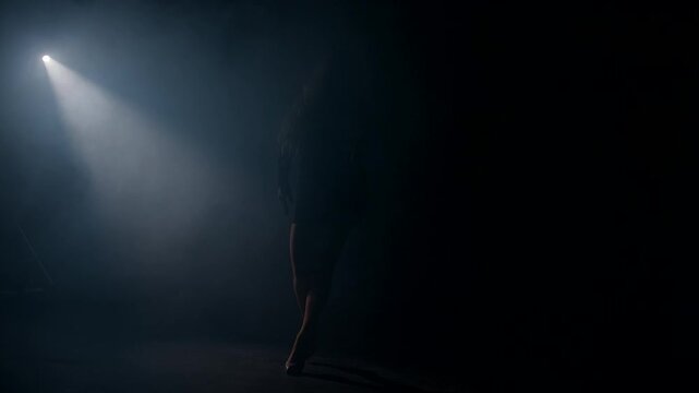 blonde woman walks confidently out of the surrounding dark space into a radiant beam of light illuminating her presence. The scene is mysterious and dramatic.