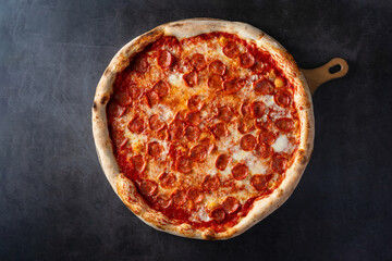 Top view of a large pizza with spicy salami on a wooden cutting board against a dark background