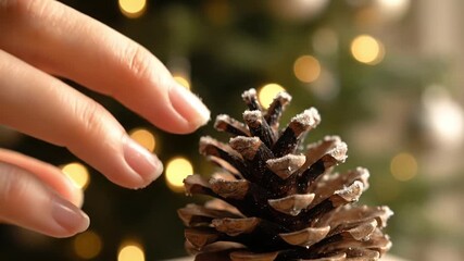 A delicate hand reaches for a frosted pine cone shimmering with ice crystals against a backdrop of soft glowing Christmas bokeh lights Ideal for winter holiday - Powered by Adobe