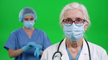 Two medical professionals an older female doctor and a younger female nurse wearing face masks and scrubs standing against a green screen ready for medical procedures or patient care. - Powered by Adobe