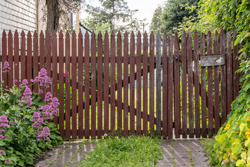 Fototapeta premium A wooden cottage-style driveway gate with vertical slats and a rustic latch, surrounded by blooming flowers. It leads into a lush, private garden in the countryside. 