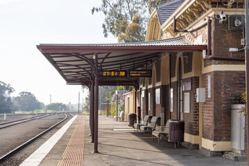 Camperdown railway station platform in regional Victoria, Australia, featuring a historic brick station building, metal canopy, seating, and an electronic timetable display. A quiet country train stop