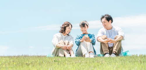A park with a view of the blue sky, a family sitting on a hill, parents and children (father, mother, daughter)

