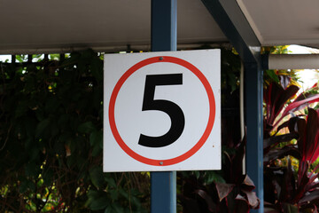 5 km/h speed limit sign mounted on a pole under a roof with tropical plants in the background