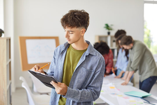 Portrait of focused young guy standing with clipboard, making notes with colleagues or classmates on background. Group of diverse people engaged in collaborative work or study project, sharing ideas.