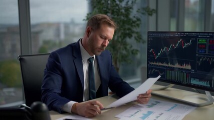 A focused businessman analyzes financial documents in his office. A monitor displays stock charts in the background, a portrait of focused attention. Stock Video - Powered by Adobe